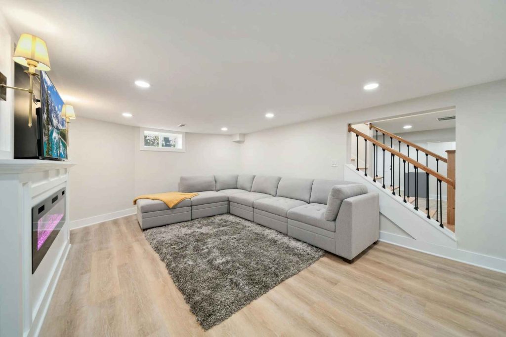 Modern basement living room with a couch, television, fireplace, and a sliding basement window letting in natural light.