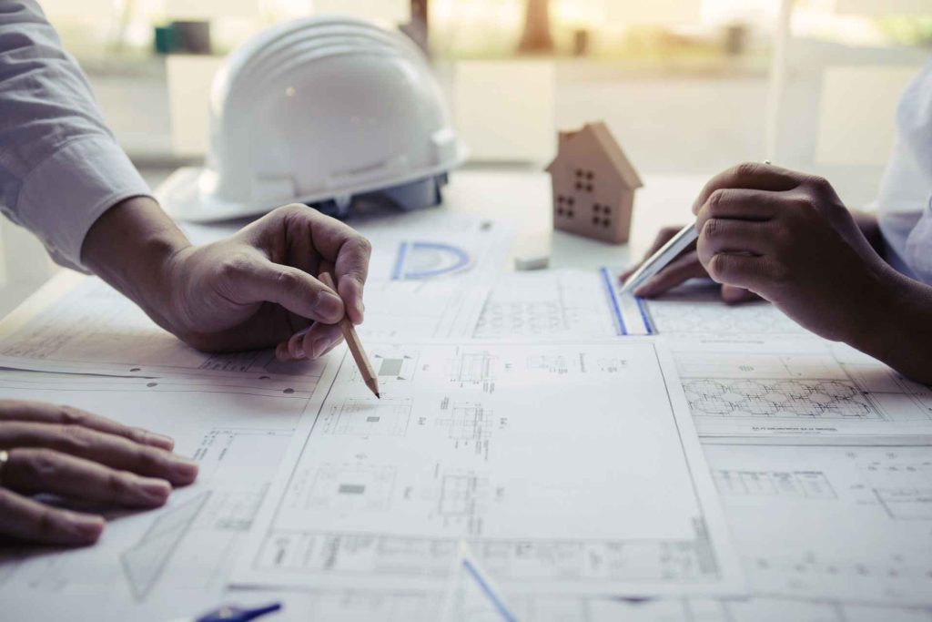 Side view of two hands working on engineering plans on a table beside a white hard hat.